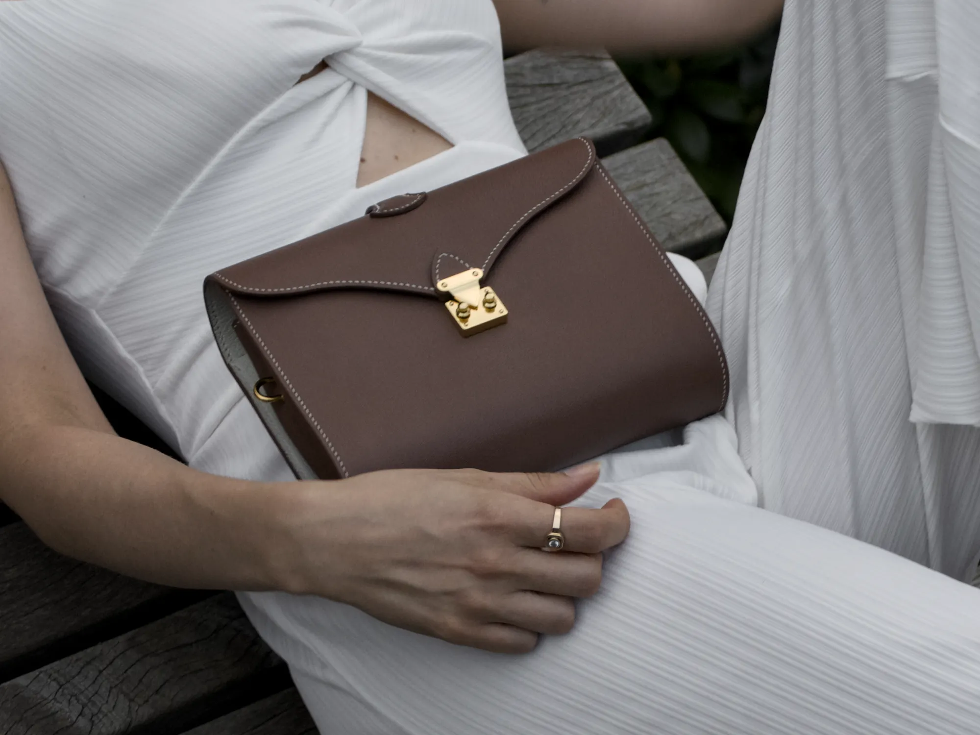 Femme assise sur un banc de pierre dans un jardin, le sac Éclat marron et crème posé contre elle, chaîne dorée