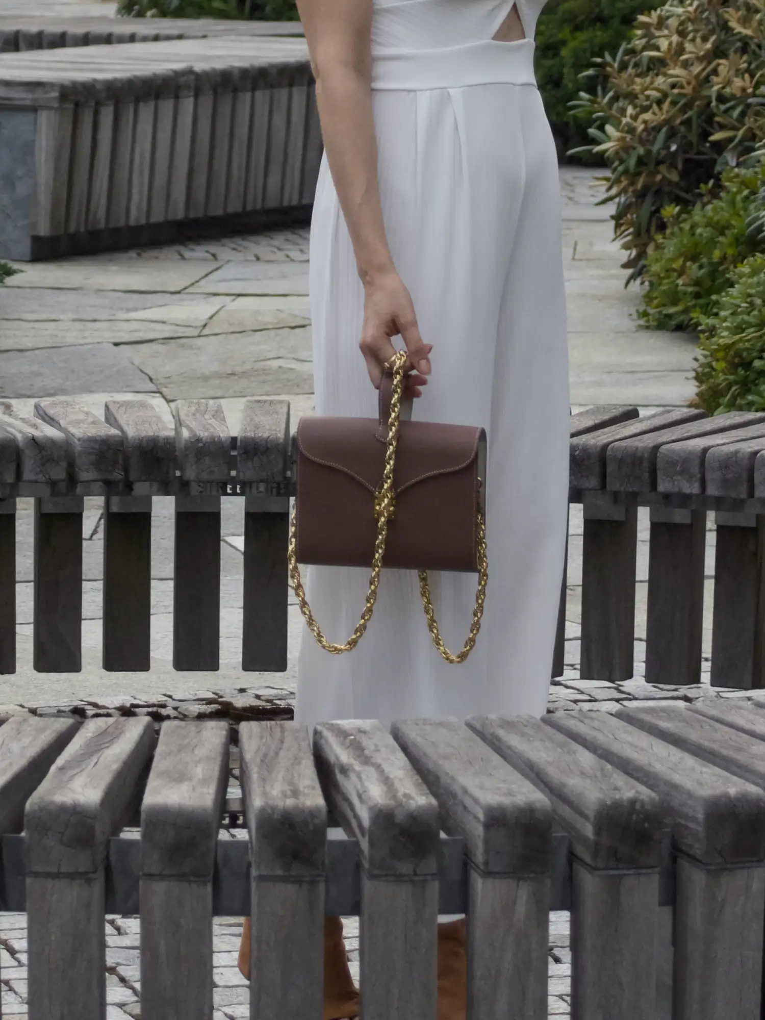 Femme de dos portant le sac Éclat marron et crème à la main, chaîne dorée visible, dans un jardin