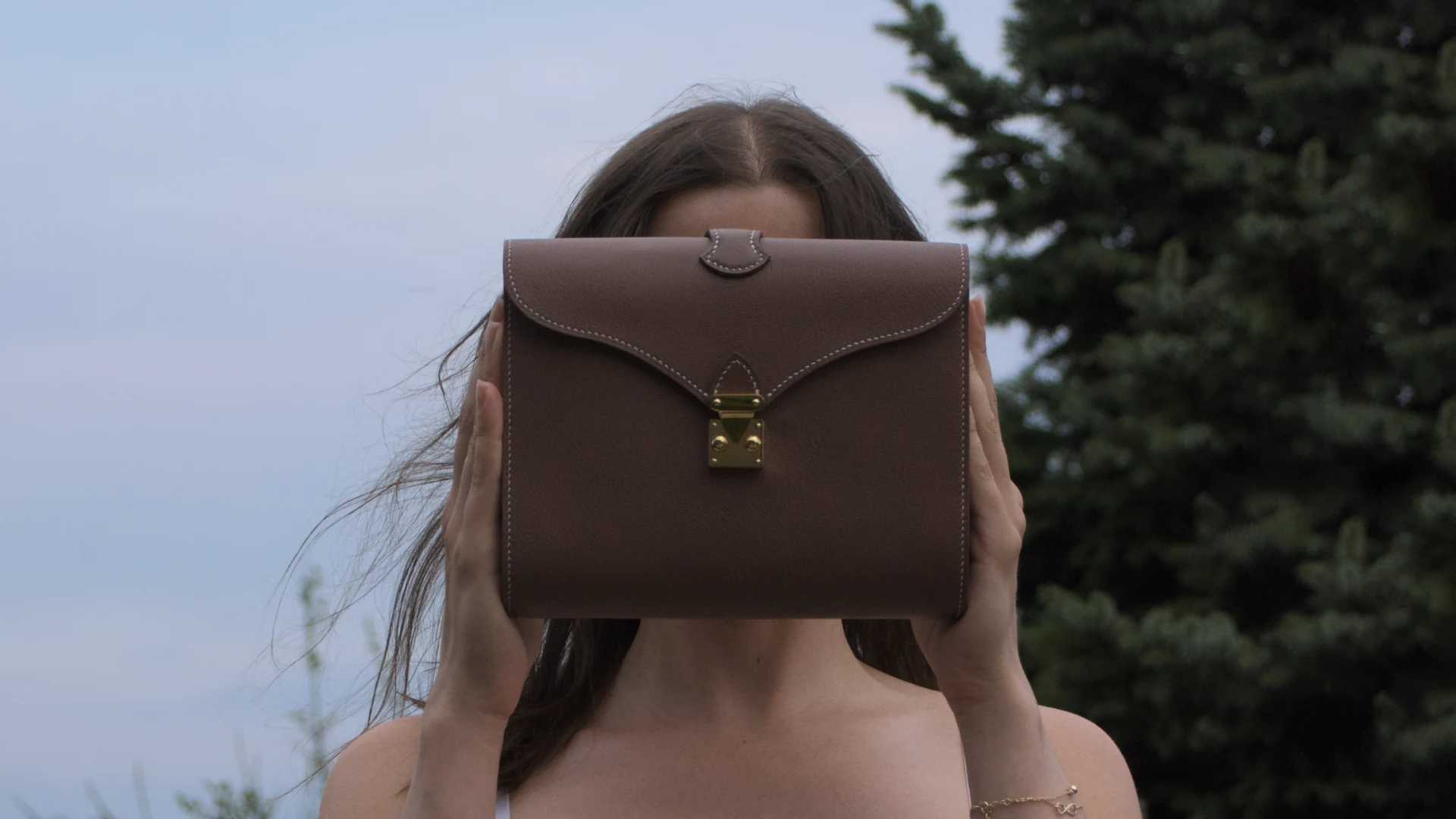 Tasche Éclat aus braunem Ziegenleder, cremefarbene Naht, goldene Beschläge, mit beiden Händen vor das Gesicht gehalten